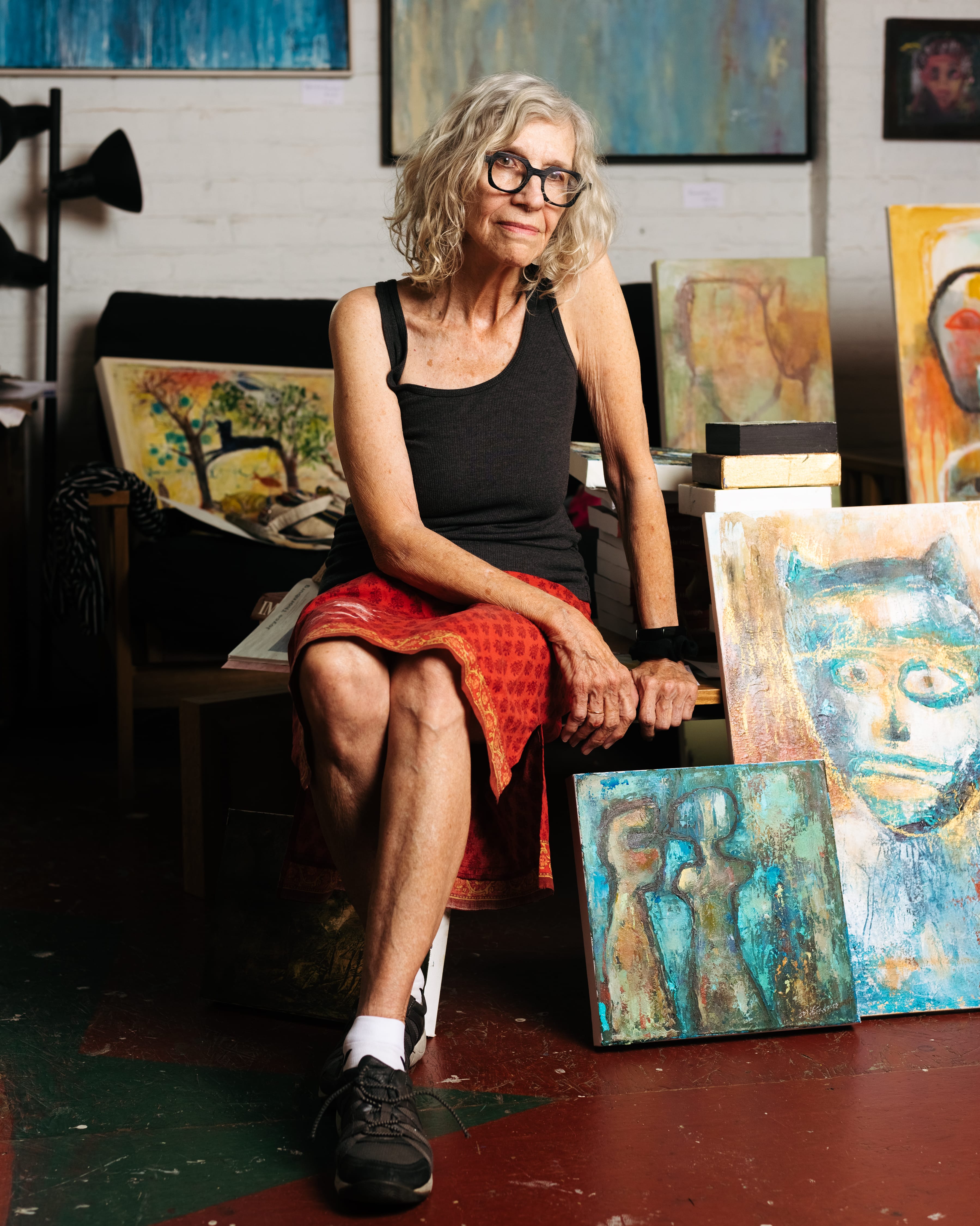 An older woman wearing thick black-framed glasses with a black tank top and red skirt sits in a chair next to paintings