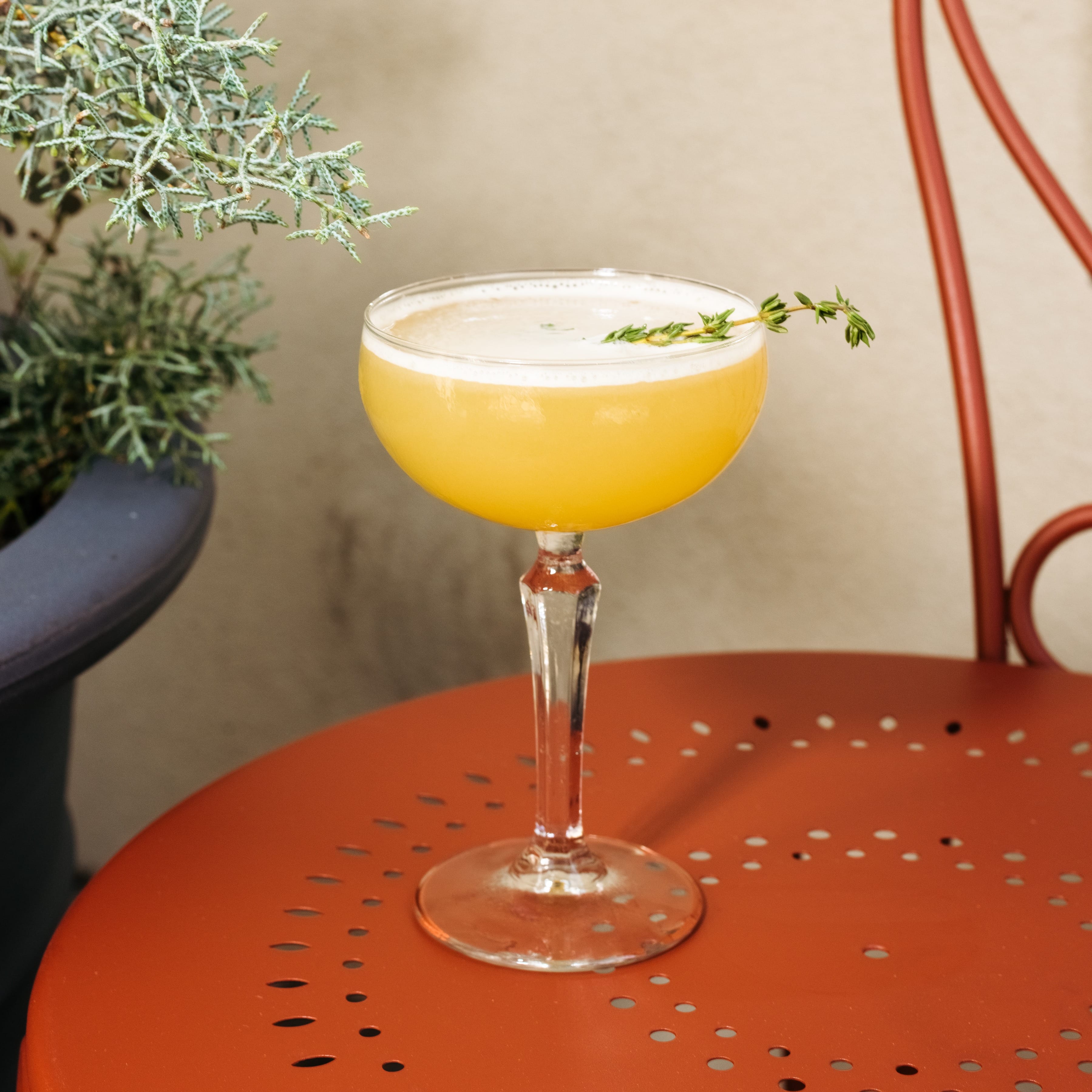 A yellow alcoholic beverage with an herbal garnish sits on an orange metal chair.