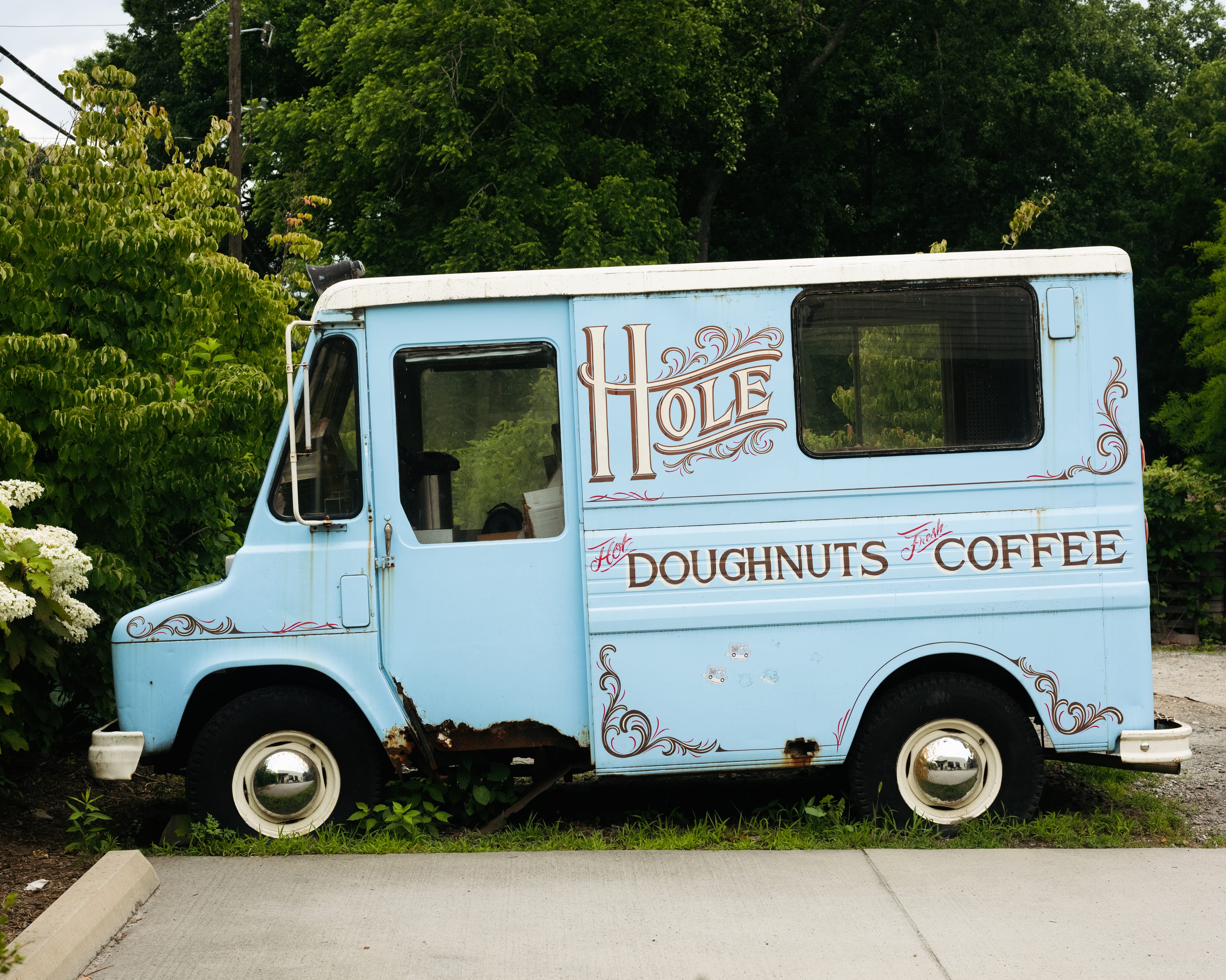 An older blue box truck with the logo for Hole Doughnuts painted on the side along with the text "Hot Doughtnuts, Fresh Coffee"